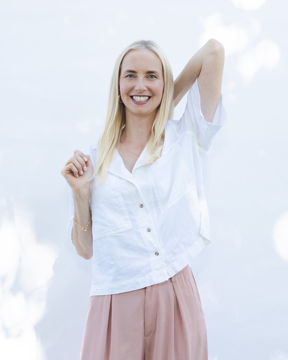 Katarina Blom ler varmt med ena armen lyft bakom huvudet, mot en ljus vägg med solfläckar